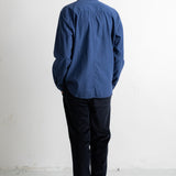 A man viewed from behind wearing a blue long-sleeve button-up shirt, dark trousers, and black-and-white sneakers, standing against a plain white wall.