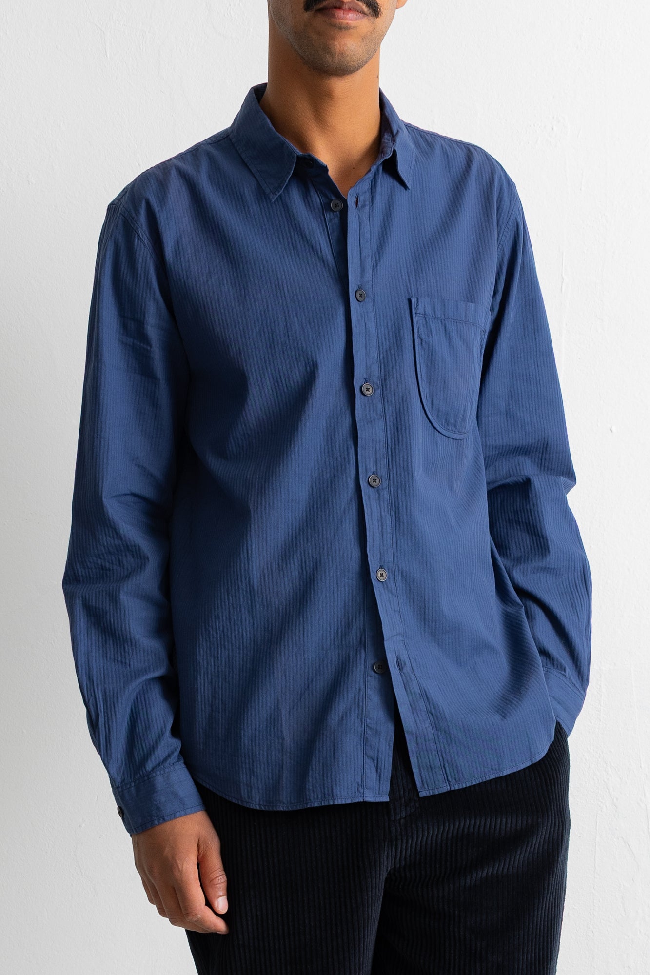 A man wearing a blue long-sleeve button-down shirt with a chest pocket, paired with dark pants, photographed against a plain light background.
