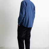 A man viewed from behind wearing a blue long-sleeve button-up shirt, dark trousers, and black canvas sneakers, standing against a plain white wall.