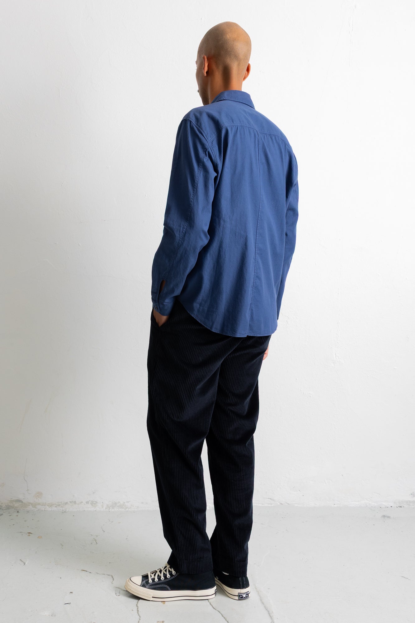 A man viewed from behind wearing a blue long-sleeve button-up shirt, dark trousers, and black canvas sneakers, standing against a plain white wall.