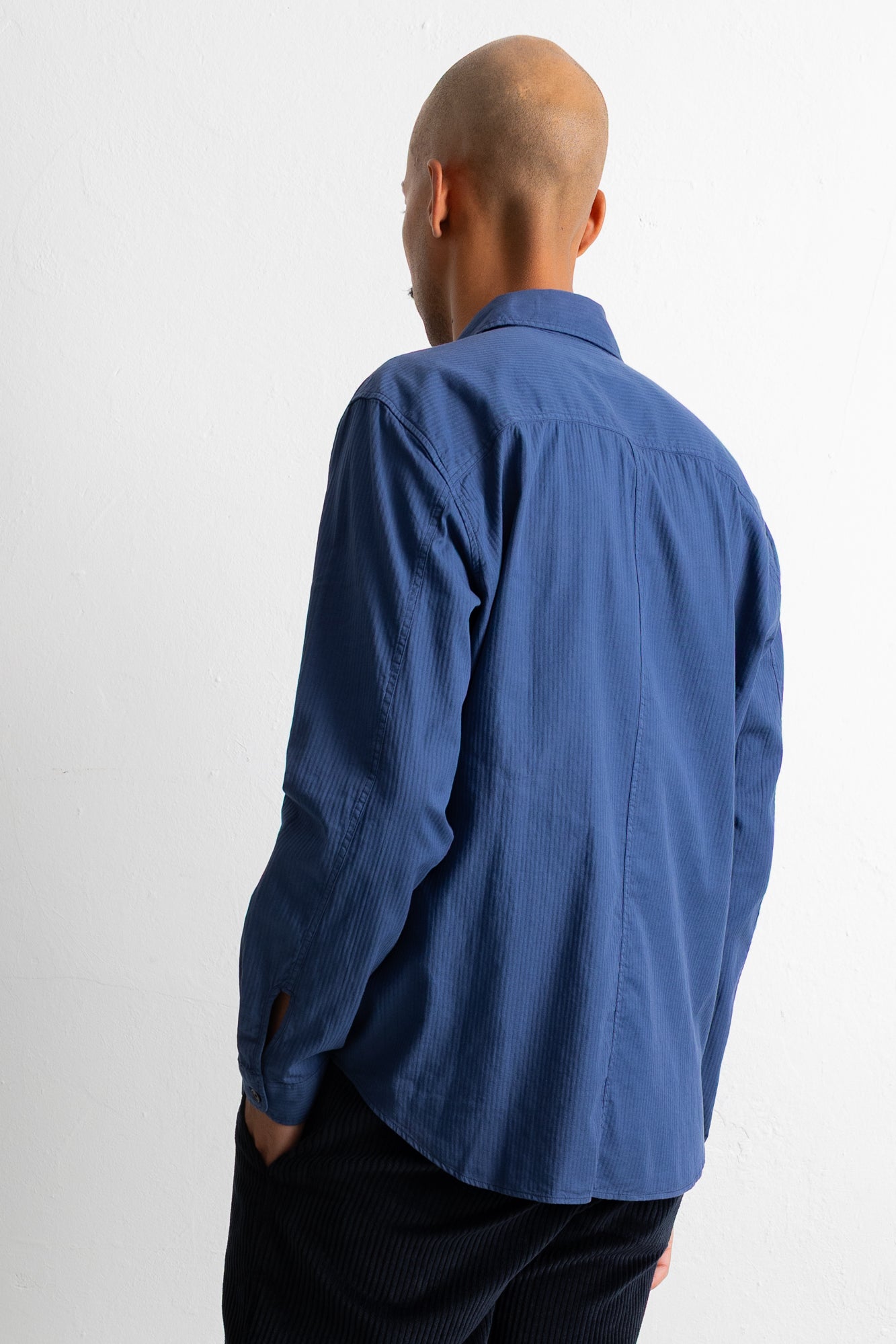 A man wearing a blue long-sleeve button-up shirt, shown from the back against a plain white studio background.
