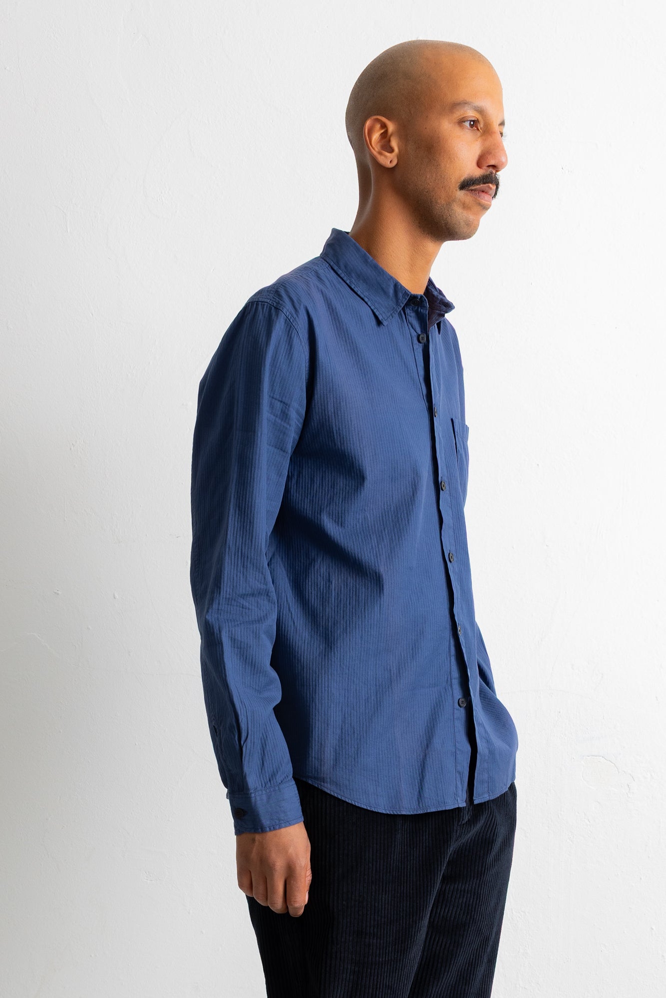 A man in profile wearing a blue long-sleeve button-down shirt and dark pants, standing against a plain light gray studio background.