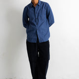 A man wearing a blue long-sleeve button-down shirt, black pants, and black canvas sneakers, standing against a plain white wall.