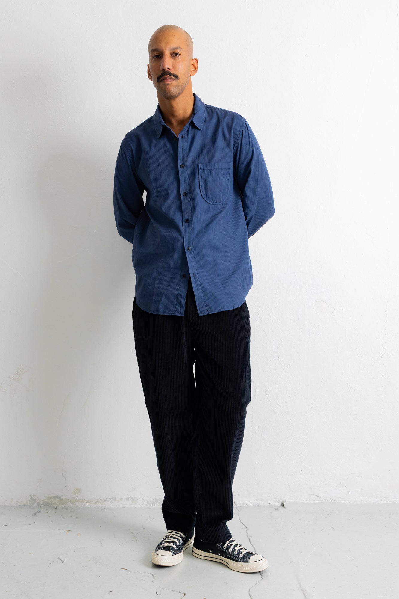 A man wearing a blue long-sleeve button-down shirt, black pants, and black canvas sneakers, standing against a plain white wall.