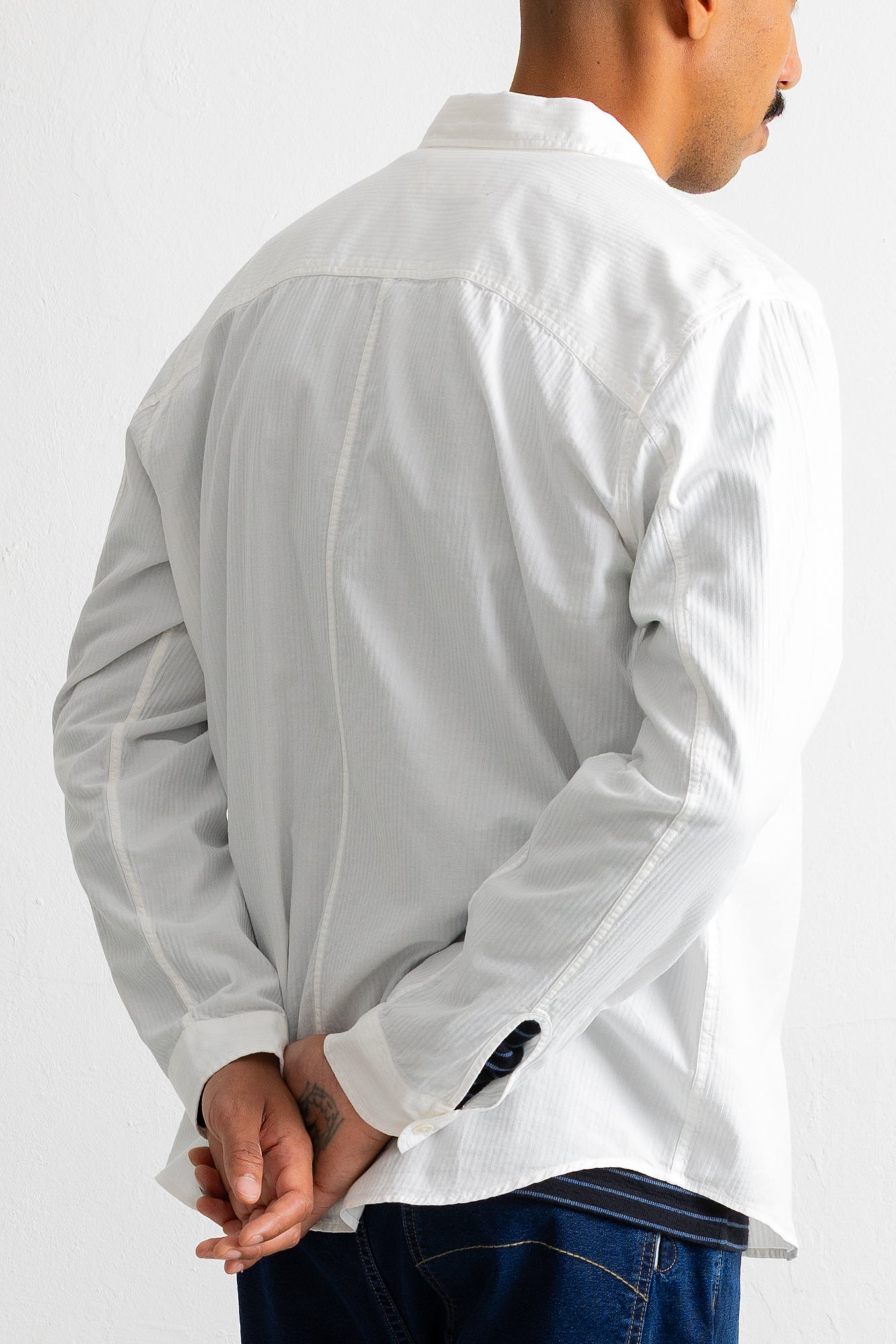 A man wearing a white long-sleeve button-up shirt, shown from the back with his hands clasped behind his back, paired with dark blue jeans against a plain white background.