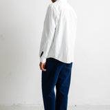 A man shown from the back wearing a white long-sleeve button-up shirt, dark navy pants, and black canvas sneakers, standing against a plain white studio wall.