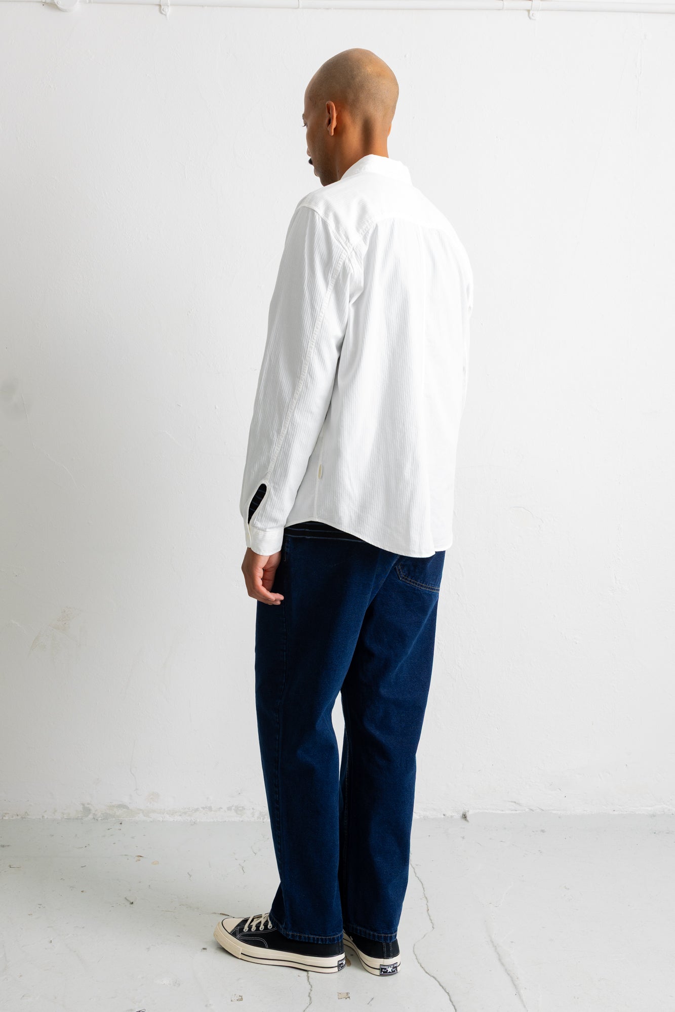 A man shown from the back wearing a white long-sleeve button-up shirt, dark navy pants, and black canvas sneakers, standing against a plain white studio wall.
