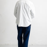 A man viewed from behind wearing a white long-sleeve button-down shirt, dark blue jeans, and black-and-white sneakers, standing against a plain white wall.