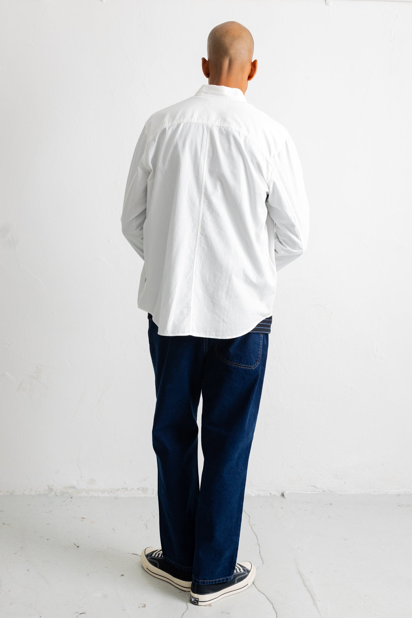 A man viewed from behind wearing a white long-sleeve button-down shirt, dark blue jeans, and black-and-white sneakers, standing against a plain white wall.