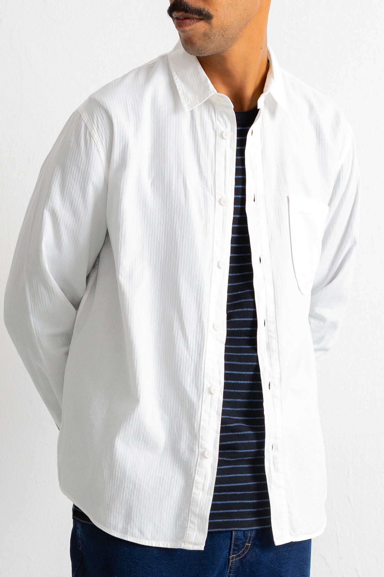 A man wearing a white long-sleeve button-down shirt over a navy blue and white striped t-shirt, paired with blue jeans, shown from the chin down against a plain white background.