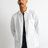 A man wearing a white long-sleeve button-up shirt over a dark striped t-shirt and blue jeans, standing against a plain white wall.