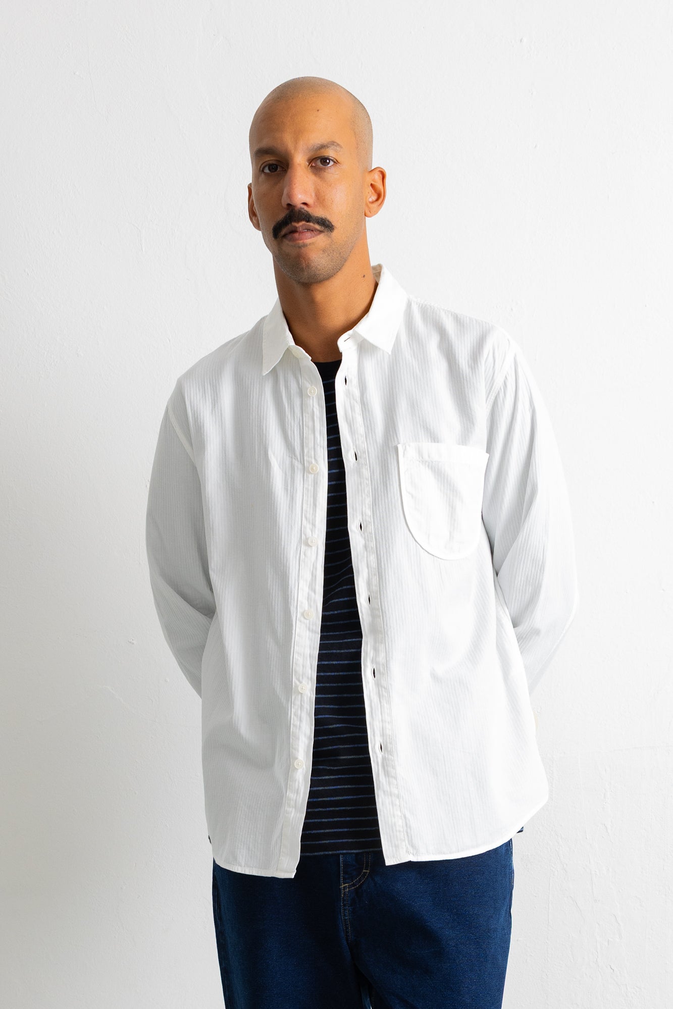 A man wearing a white long-sleeve button-up shirt over a dark striped t-shirt and blue jeans, standing against a plain white wall.