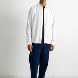 A man wearing a white long-sleeve button-up shirt over a black and white striped shirt, paired with dark blue trousers and black-and-white sneakers, standing against a plain white wall.