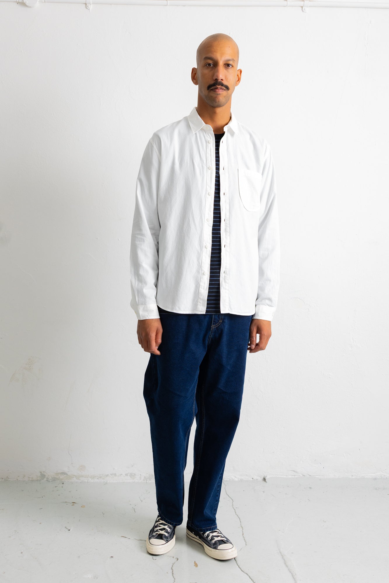 A man wearing a white long-sleeve button-up shirt over a black and white striped shirt, paired with dark blue trousers and black-and-white sneakers, standing against a plain white wall.