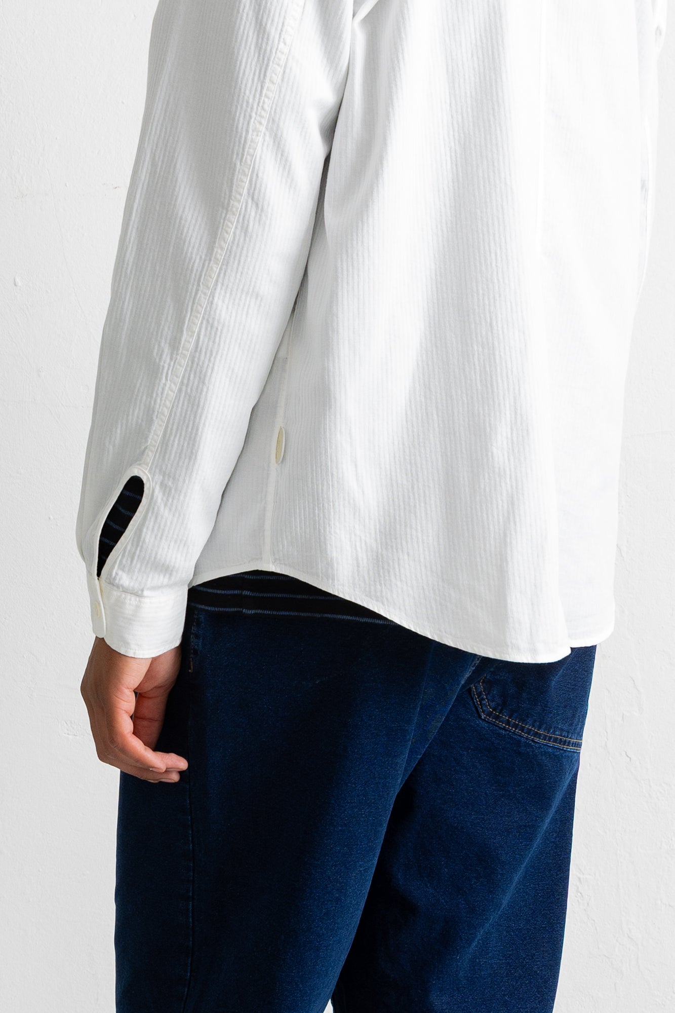 Close-up of a person wearing a white textured long-sleeve button-up shirt and dark blue jeans, showing the back of the shirt and sleeve detail against a plain light background.