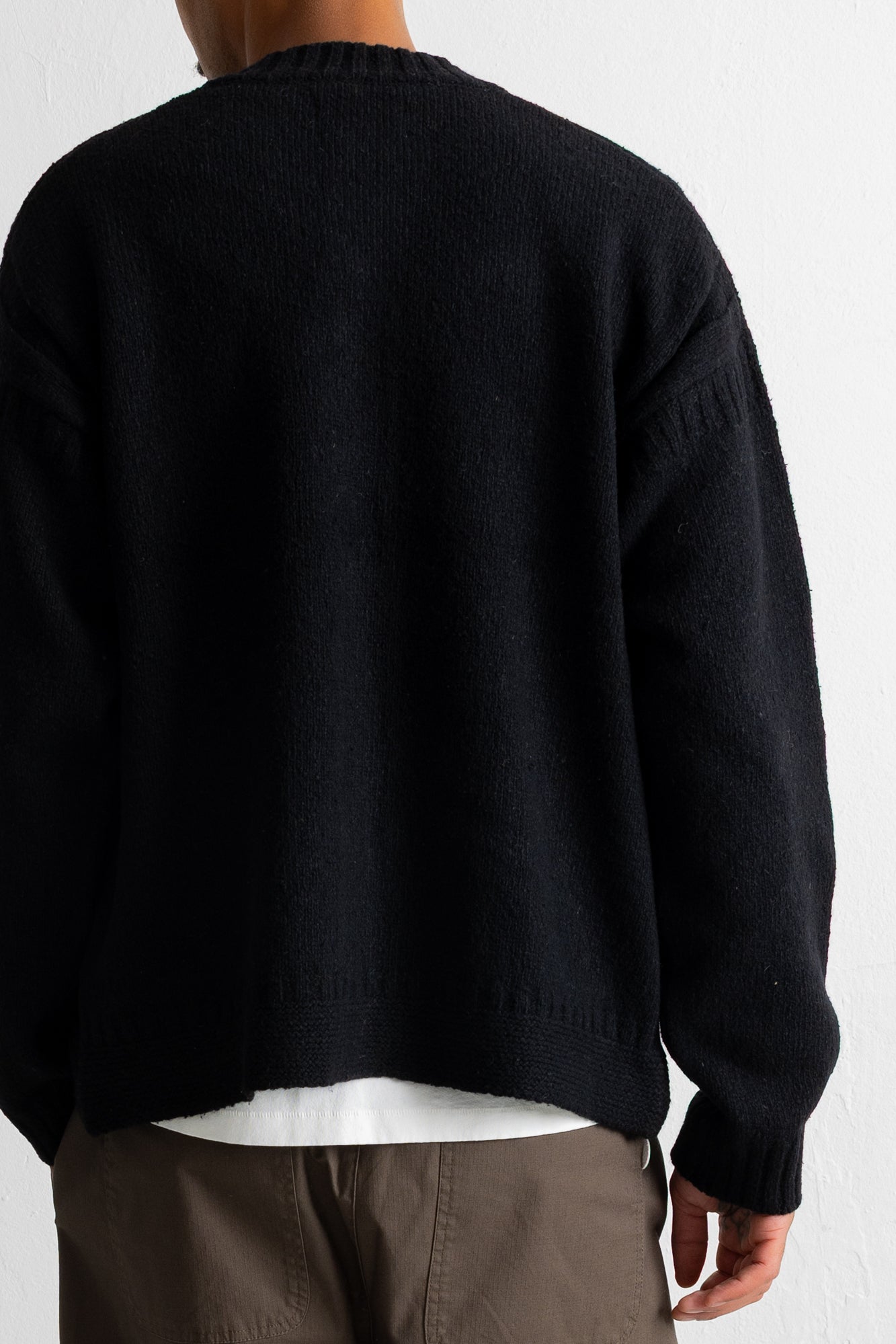 A person shown from the back wearing a black knit sweater over a white undershirt and brown pants, photographed against a plain light background.