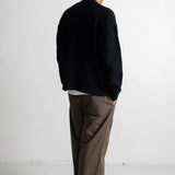 A man viewed from behind wearing a black knit sweater, brown wide-leg trousers, and black canvas sneakers, standing against a white wall.