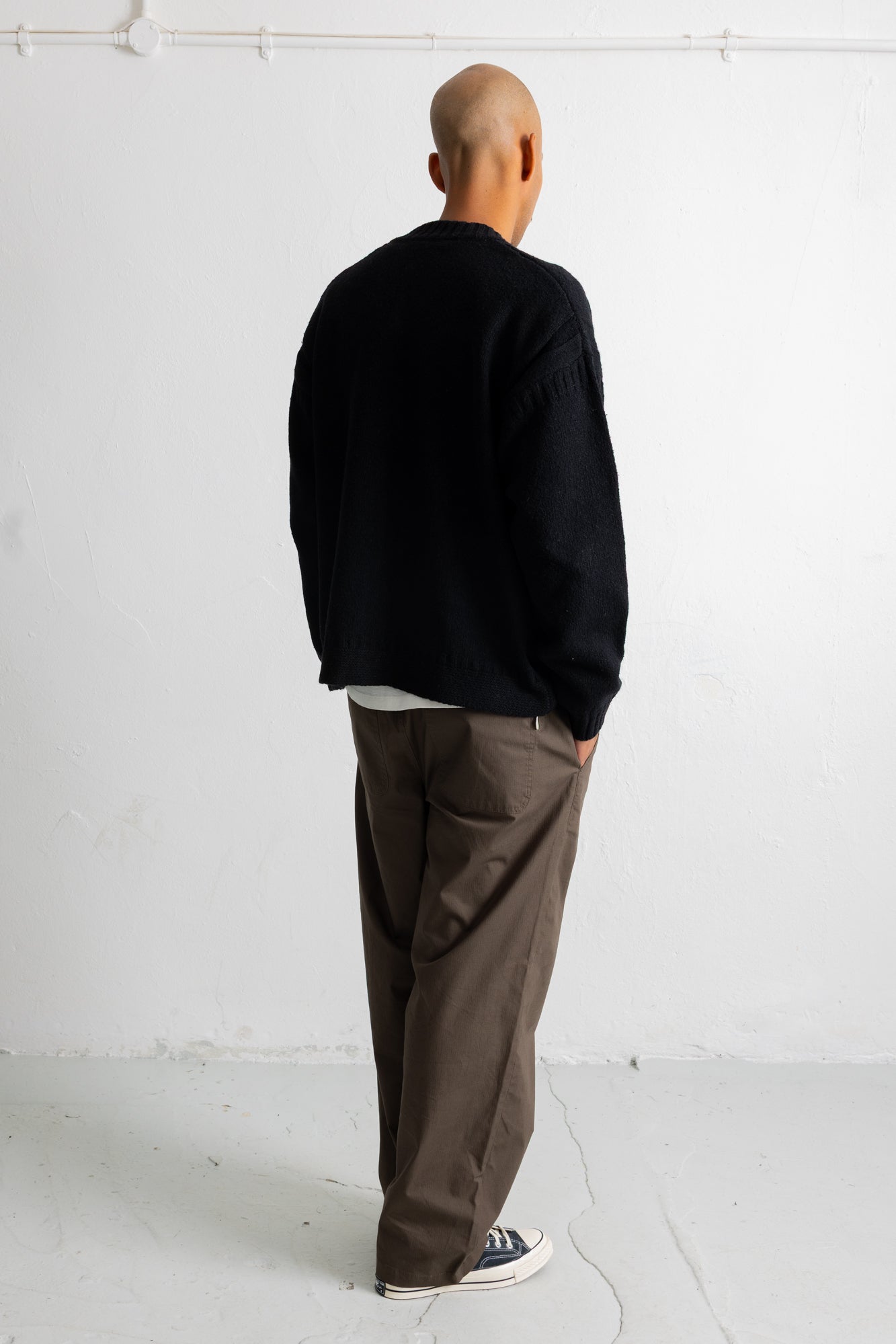 A man viewed from behind wearing a black knit sweater, brown wide-leg trousers, and black canvas sneakers, standing against a white wall.