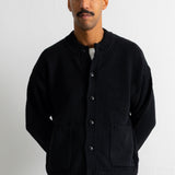 A man wearing a black knit button-up cardigan with front pockets, styled over a white undershirt and brown trousers, standing against a plain light gray wall.