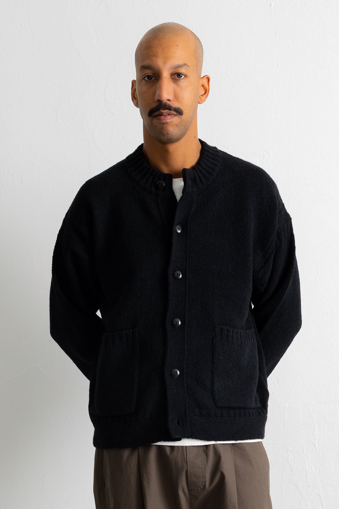A man wearing a black knit button-up cardigan with front pockets, styled over a white undershirt and brown trousers, standing against a plain light gray wall.