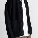 A man wearing a black knit cardigan over a white t-shirt and brown trousers, shown from the neck down against a light gray wall.