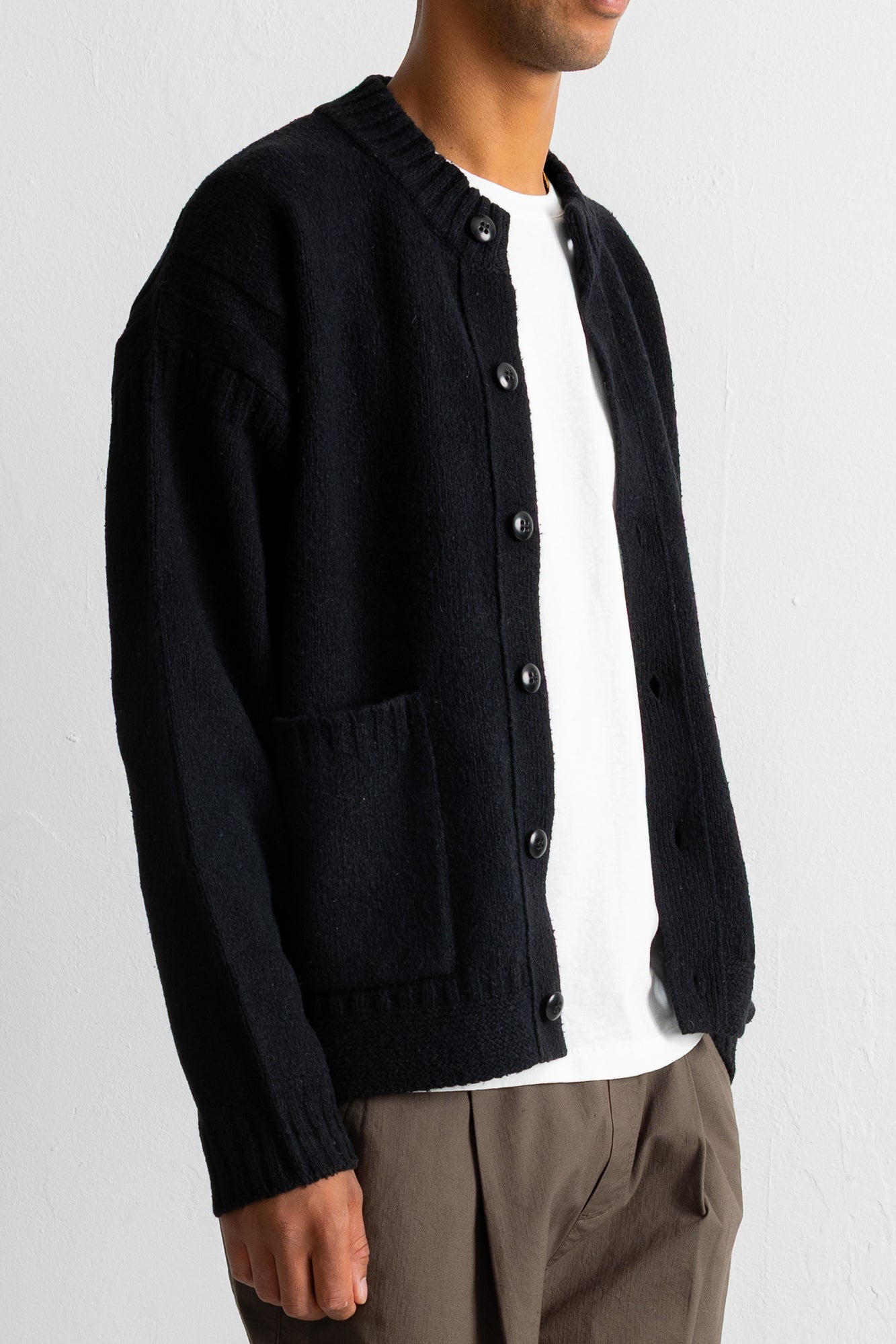 A man wearing a black knit cardigan over a white t-shirt and brown trousers, shown from the neck down against a light gray wall.