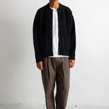 A man wearing a black corduroy button-up jacket over a white t-shirt, paired with brown trousers and black-and-white sneakers, standing against a white studio wall.