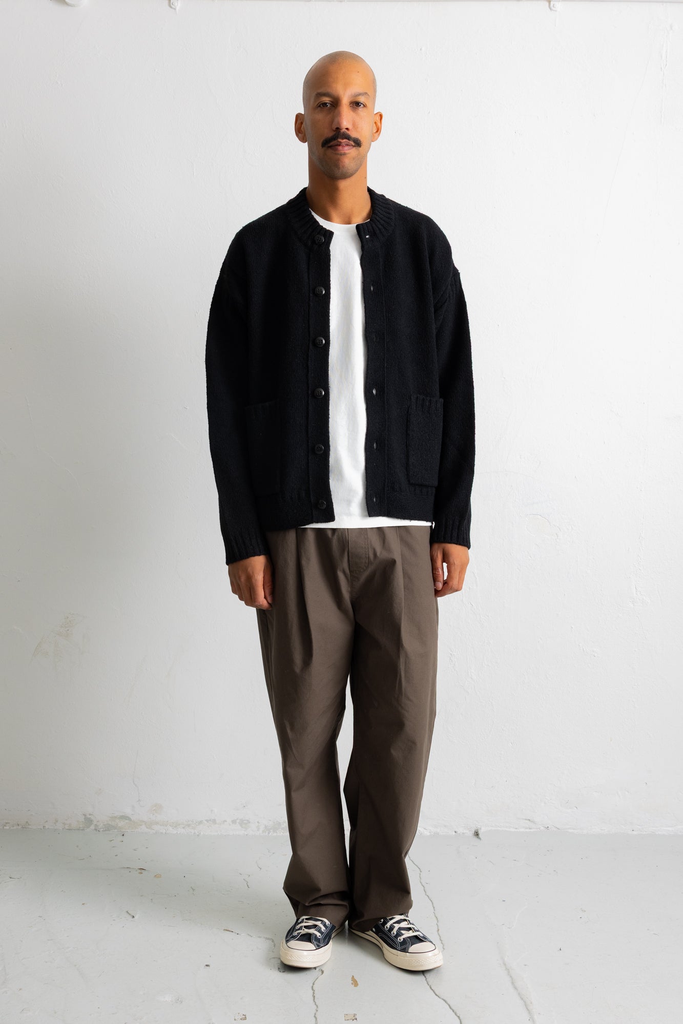 A man wearing a black corduroy button-up jacket over a white t-shirt, paired with brown trousers and black-and-white sneakers, standing against a white studio wall.