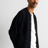 A man wearing a black knit button-up cardigan over a white t-shirt and brown trousers, photographed against a plain light gray background.