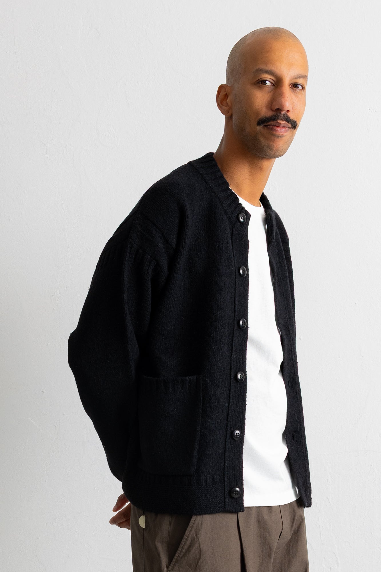 A man wearing a black knit button-up cardigan over a white t-shirt and brown trousers, photographed against a plain light gray background.
