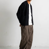 A man wearing a black corduroy button-up cardigan over a white t-shirt, paired with brown wide-leg trousers and black canvas sneakers, standing against a white studio wall.