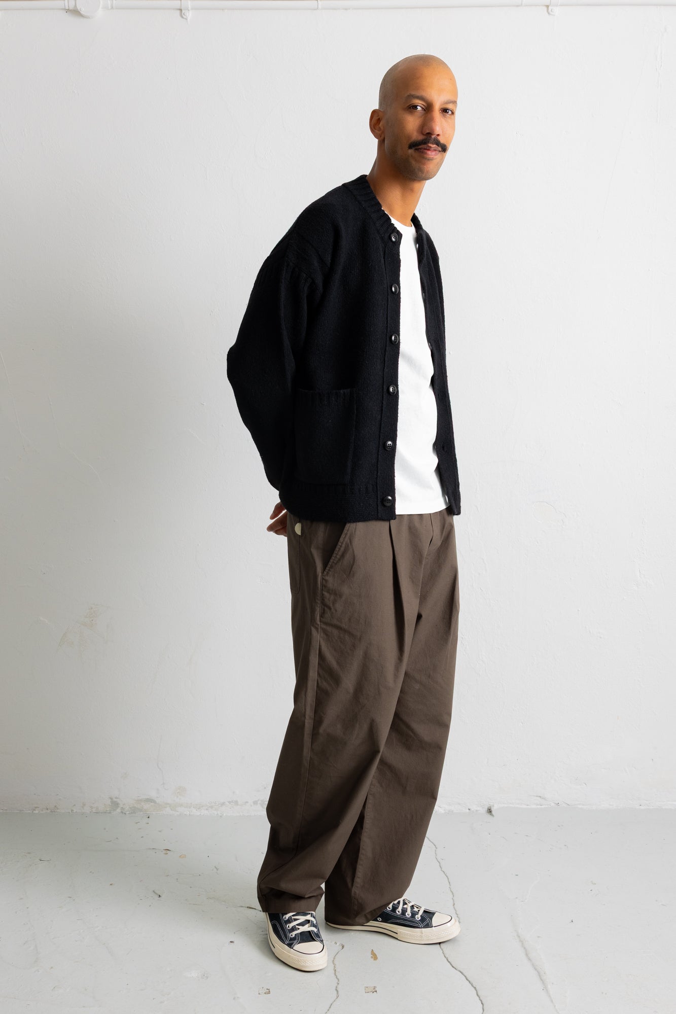 A man wearing a black corduroy button-up cardigan over a white t-shirt, paired with brown wide-leg trousers and black canvas sneakers, standing against a white studio wall.