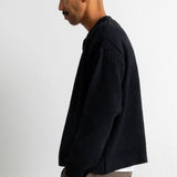 A man in profile wearing a black knit sweater and taupe trousers, standing against a plain white wall.