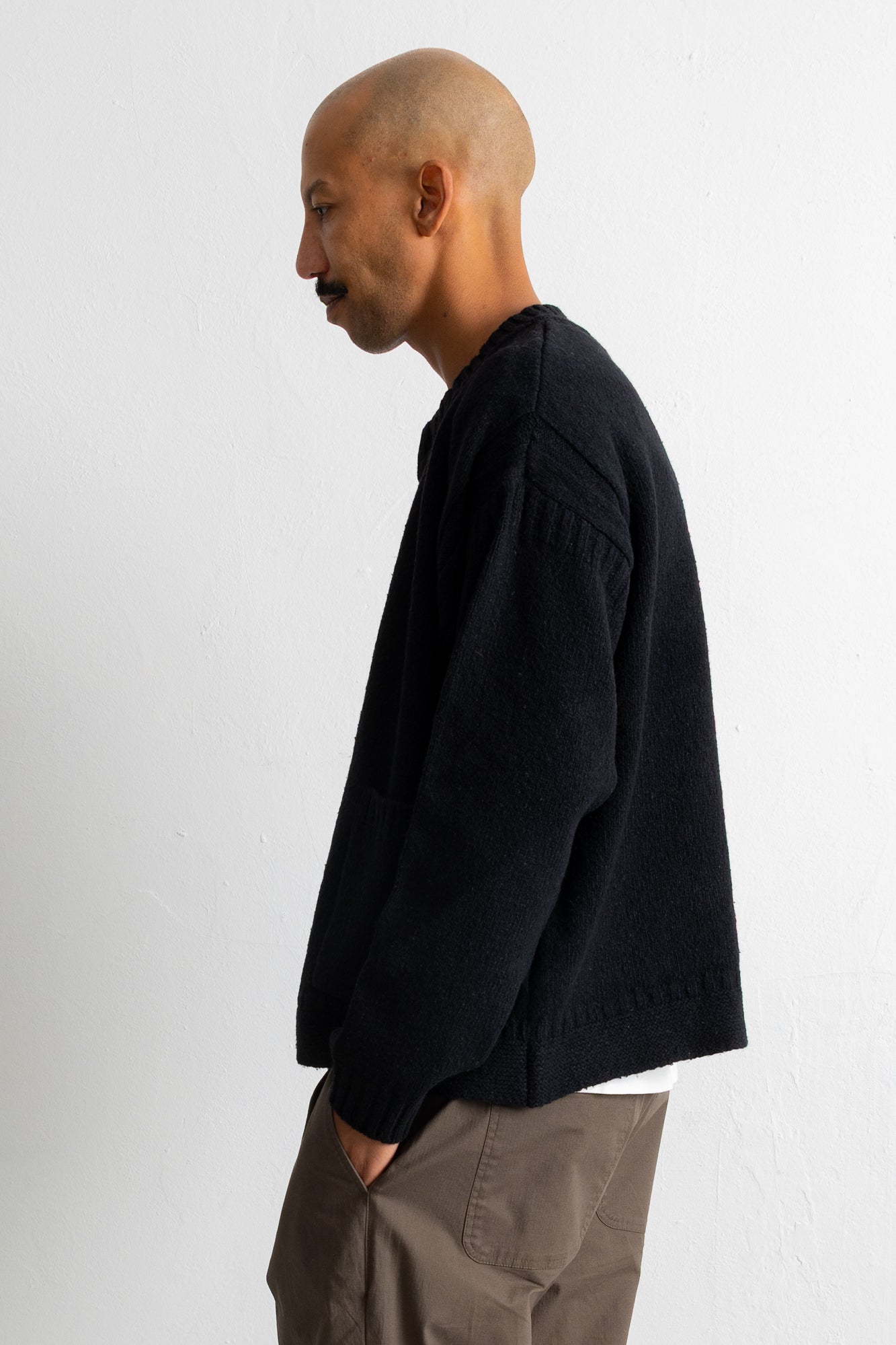 A man in profile wearing a black knit sweater and taupe trousers, standing against a plain white wall.