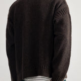 A man viewed from the back wearing a dark brown knit sweater, a white and black striped t-shirt, and blue jeans against a plain light gray background.