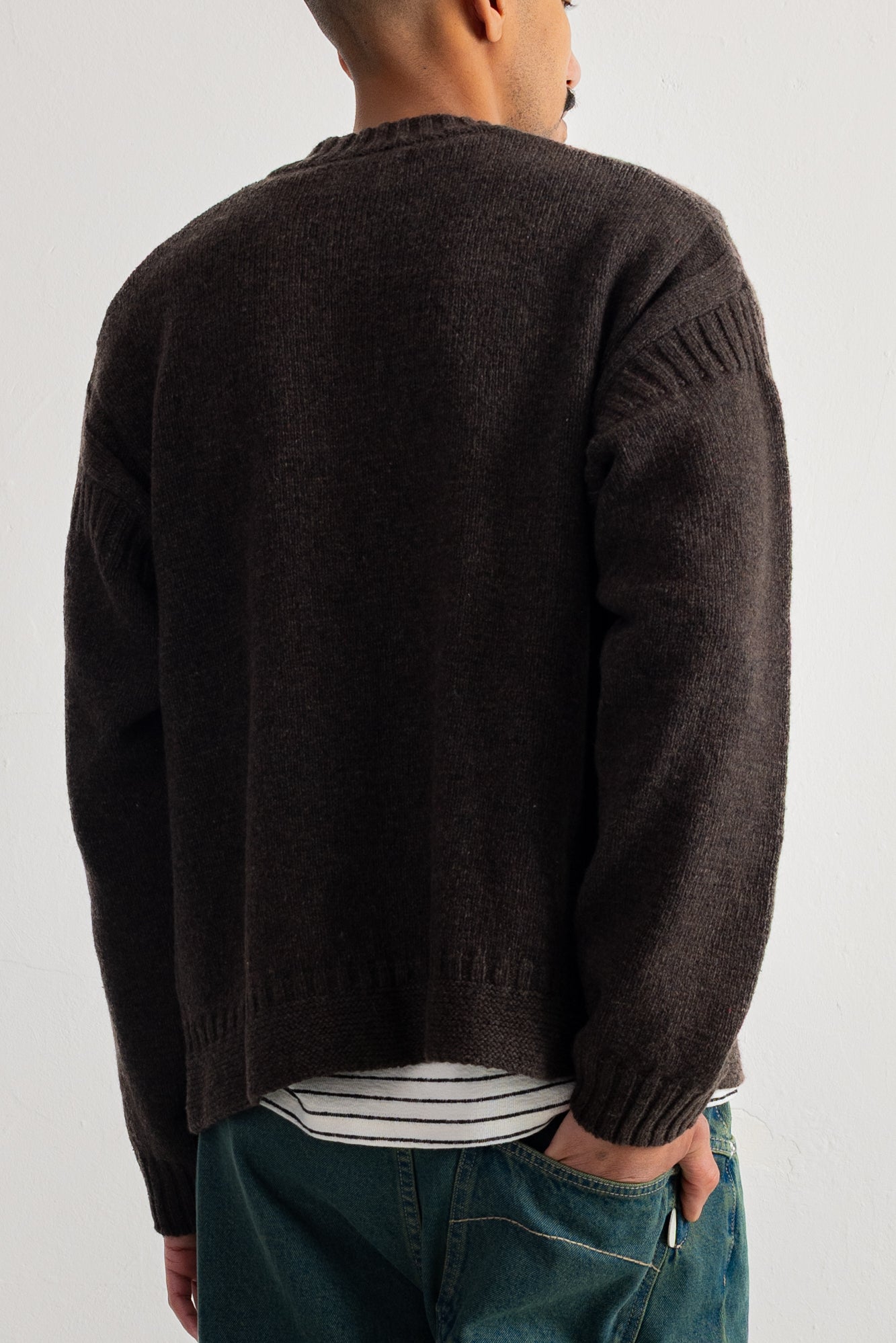 A man viewed from the back wearing a dark brown knit sweater, a white and black striped t-shirt, and blue jeans against a plain light gray background.