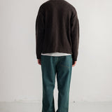 A man viewed from behind wearing a dark brown knit sweater, a white t-shirt, dark green trousers, and black shoes, standing against a plain white studio wall.