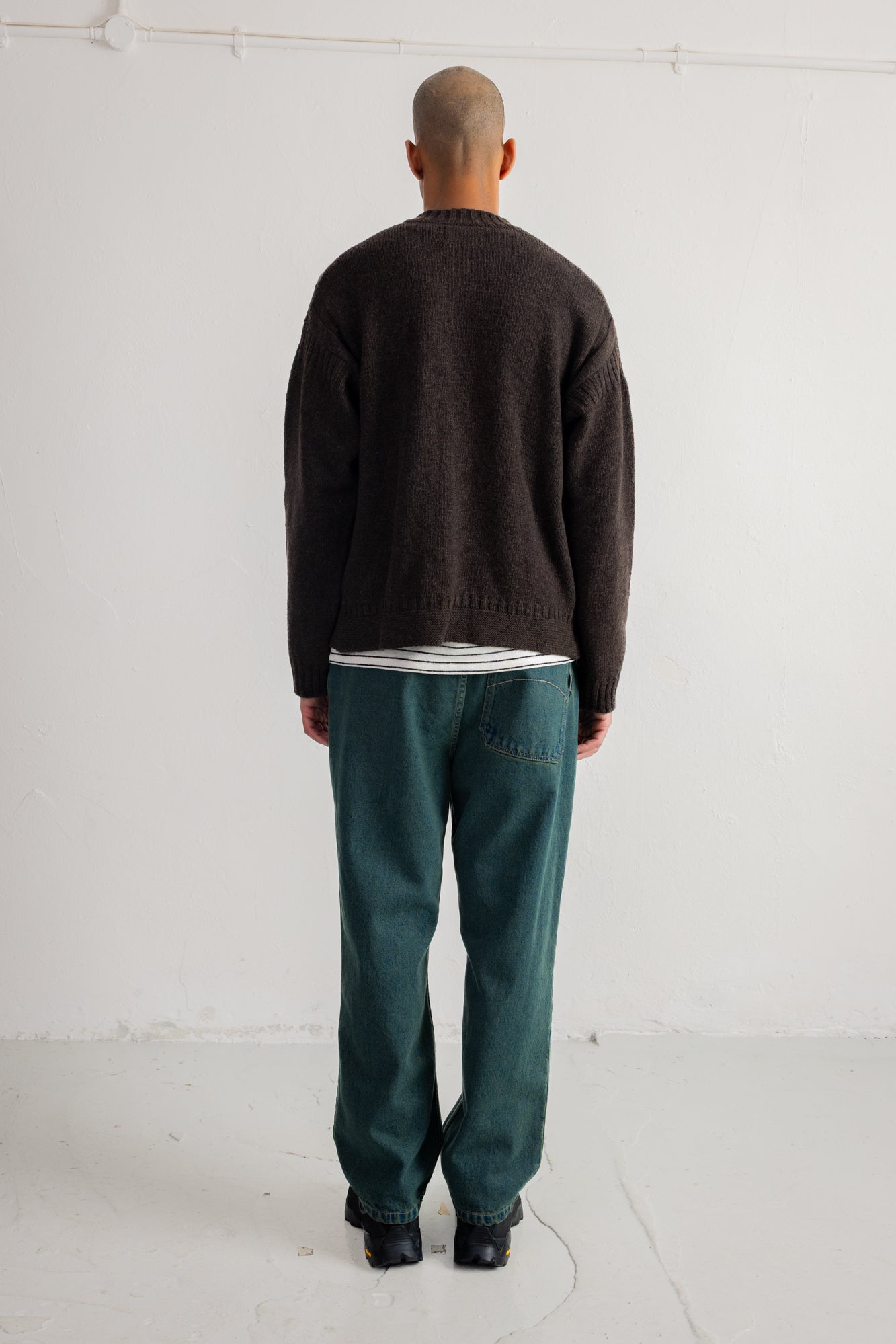 A man viewed from behind wearing a dark brown knit sweater, a white t-shirt, dark green trousers, and black shoes, standing against a plain white studio wall.