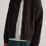 A man wearing a dark brown knit cardigan over a white and black striped t-shirt, paired with teal jeans, photographed against a light gray studio background.