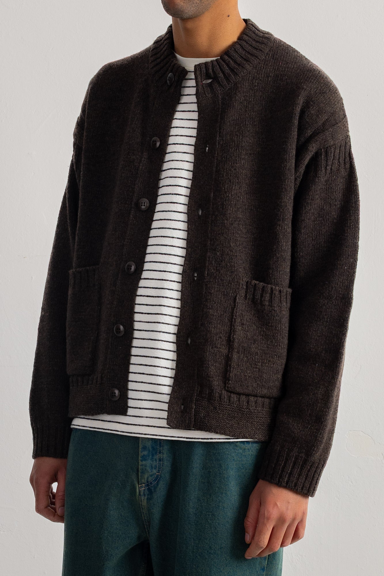 A man wearing a dark brown knit cardigan over a white and black striped t-shirt, paired with teal jeans, photographed against a light gray studio background.