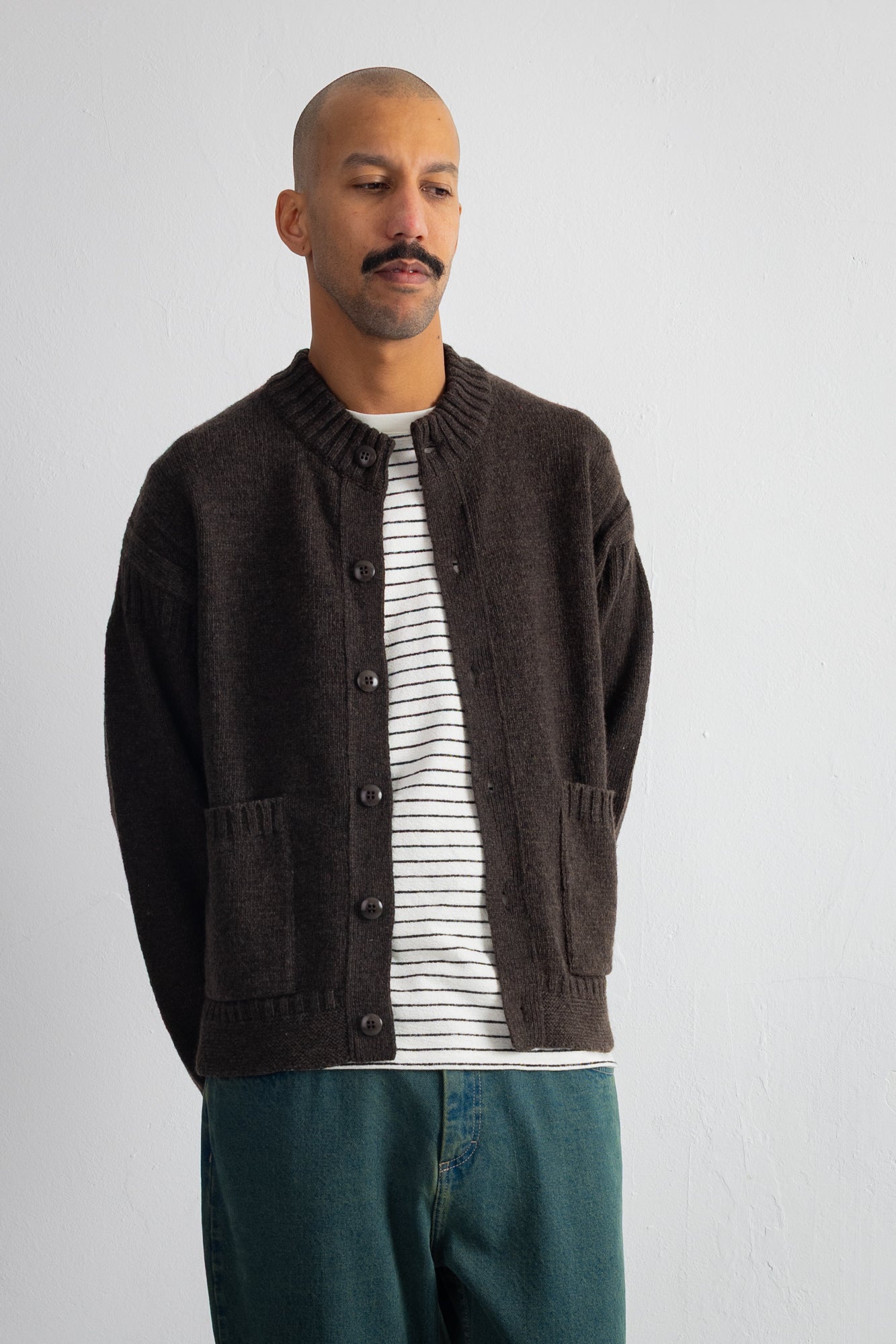A man wearing a dark brown knit button-up cardigan over a white and black striped t-shirt, paired with teal jeans, standing against a light gray wall.