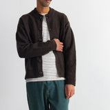 A man wearing a dark brown knit cardigan over a white and gray striped t-shirt, paired with teal jeans, standing against a plain light gray wall.