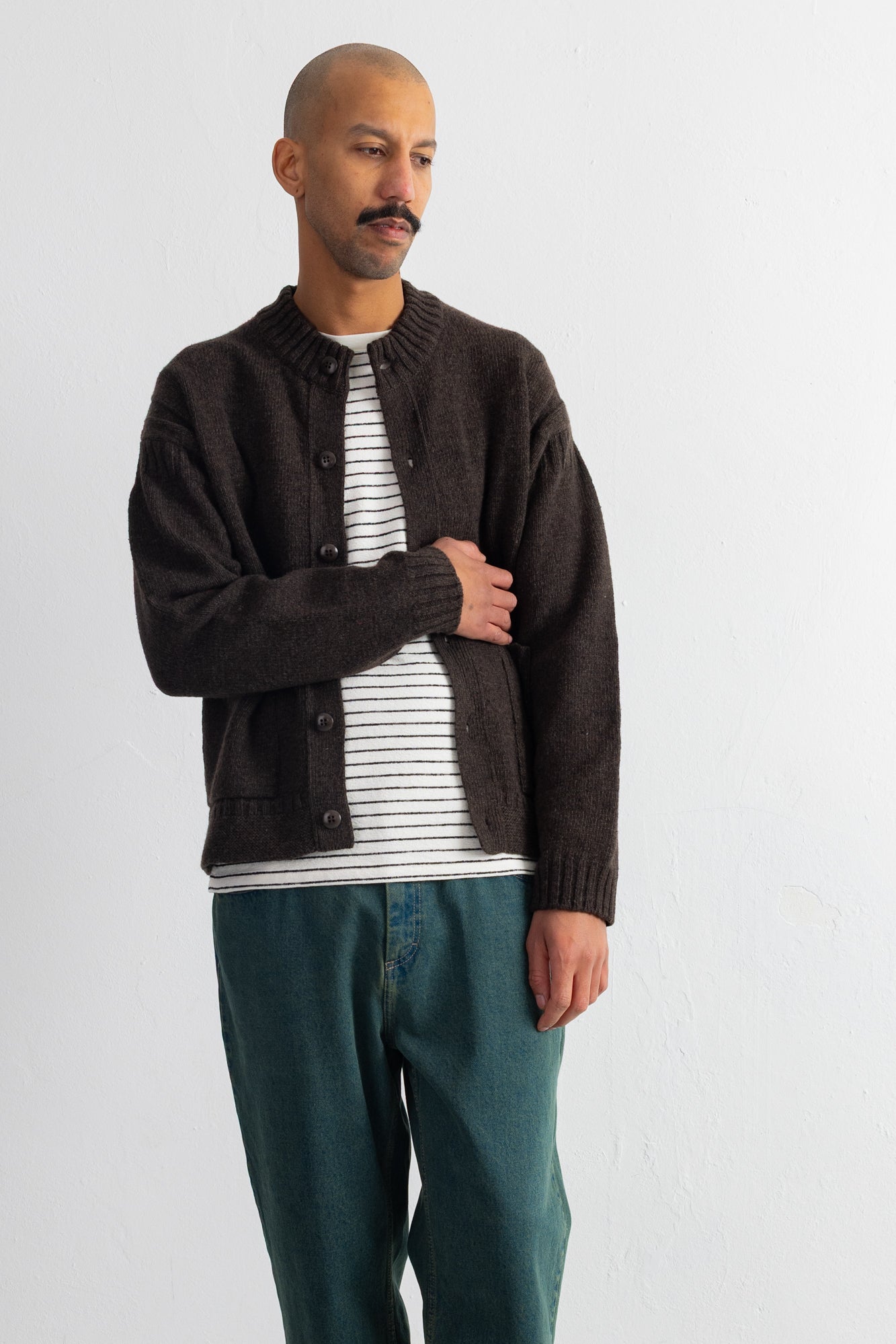 A man wearing a dark brown knit cardigan over a white and gray striped t-shirt, paired with teal jeans, standing against a plain light gray wall.