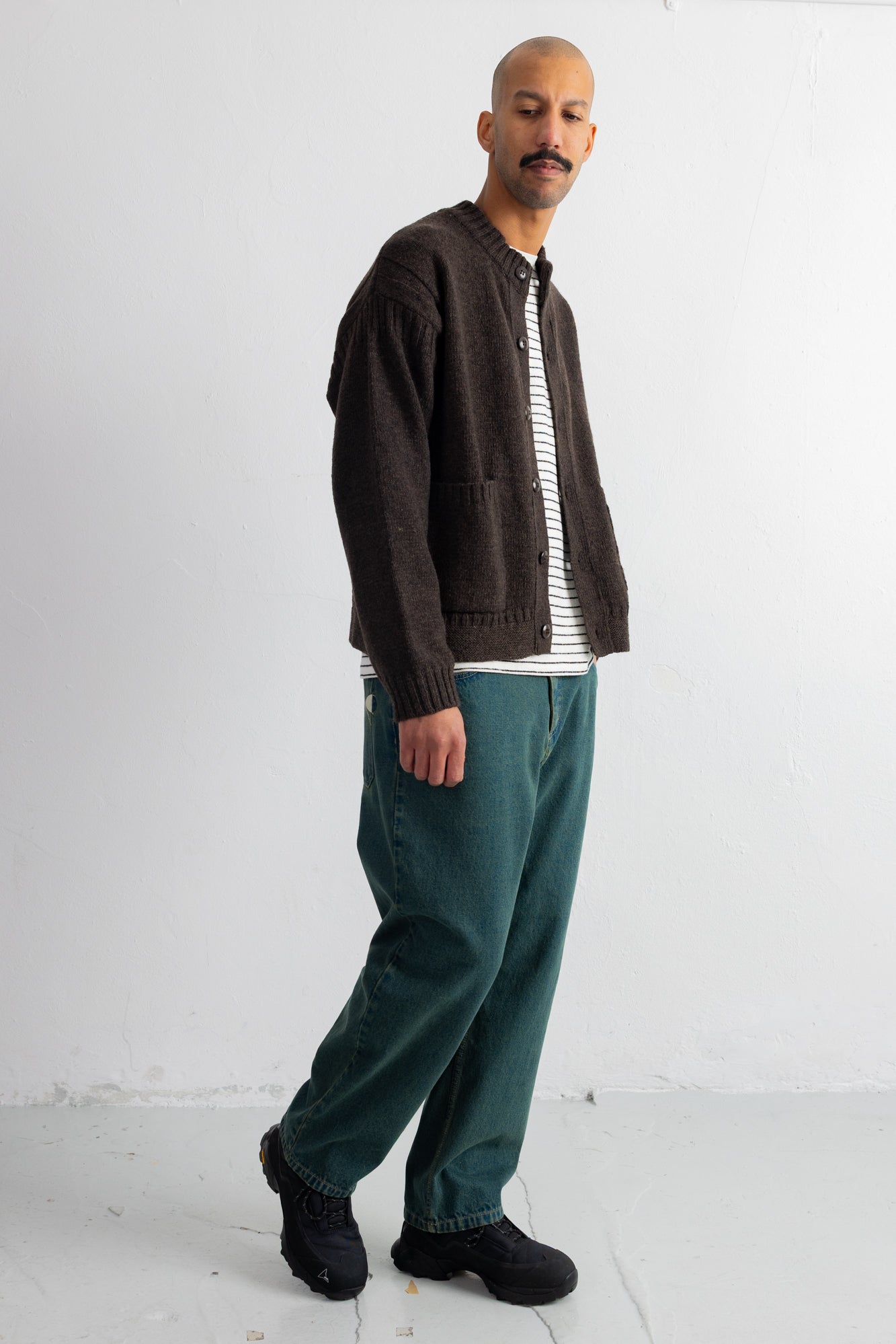A man wearing a dark brown knit cardigan over a black and white striped shirt, paired with teal green trousers and black shoes, standing against a plain white studio background.