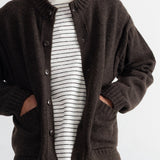 A man wearing a dark brown knit cardigan over a white and black striped t-shirt and blue jeans, shown from the neck down against a plain light background.