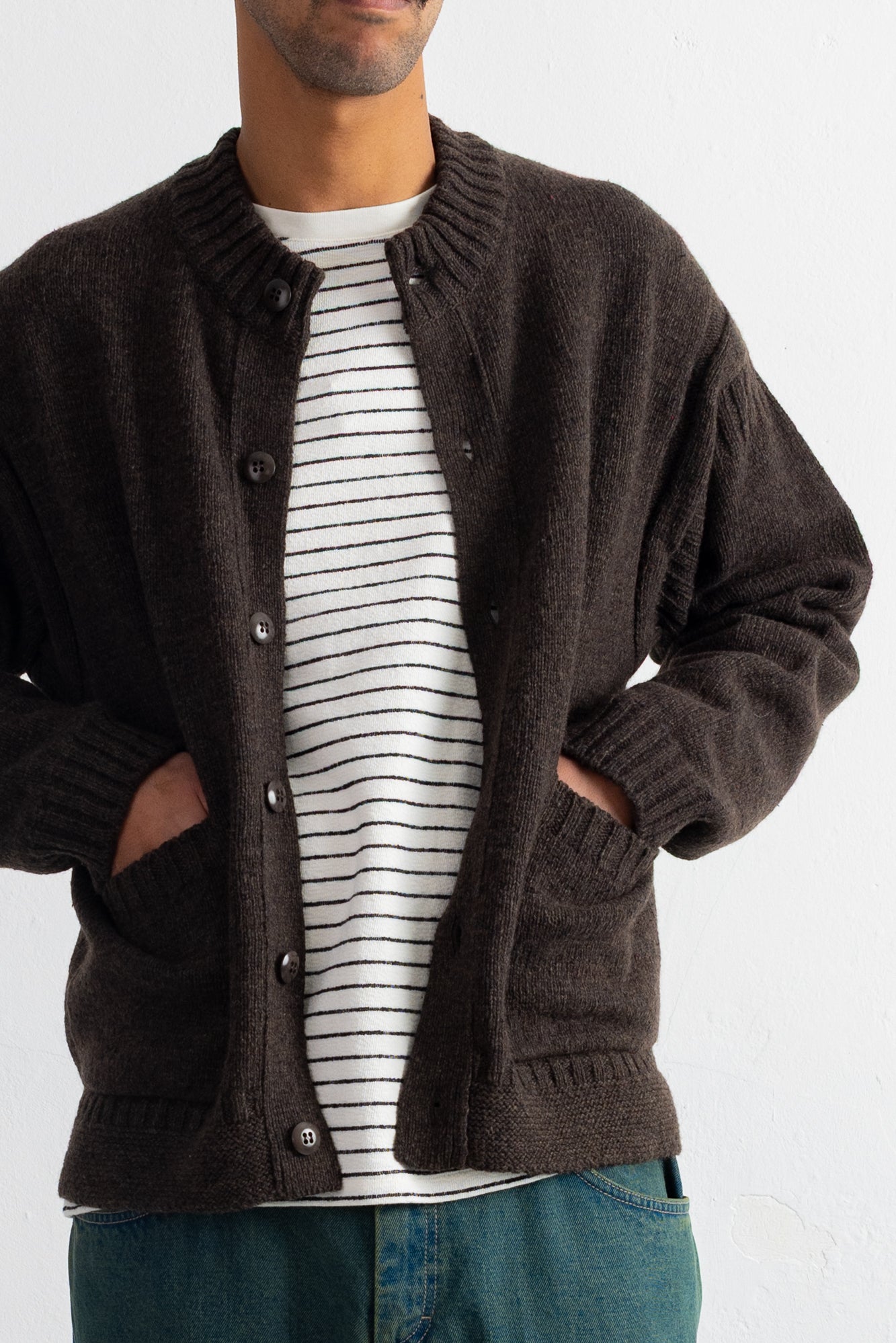 A man wearing a dark brown knit cardigan over a white and black striped t-shirt and blue jeans, shown from the neck down against a plain light background.