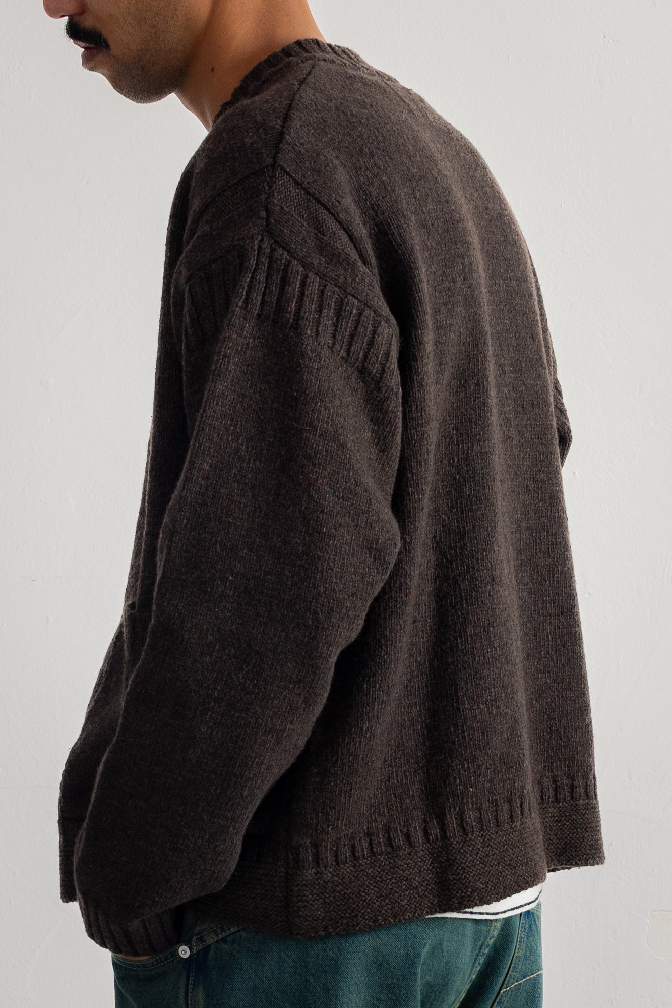 A man wearing a dark brown knit sweater, shown from a side-back angle against a plain light gray background.