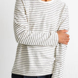 A man wearing a long-sleeve crewneck shirt with thin black horizontal stripes on an off-white base, paired with dark blue pants, against a plain white wall.