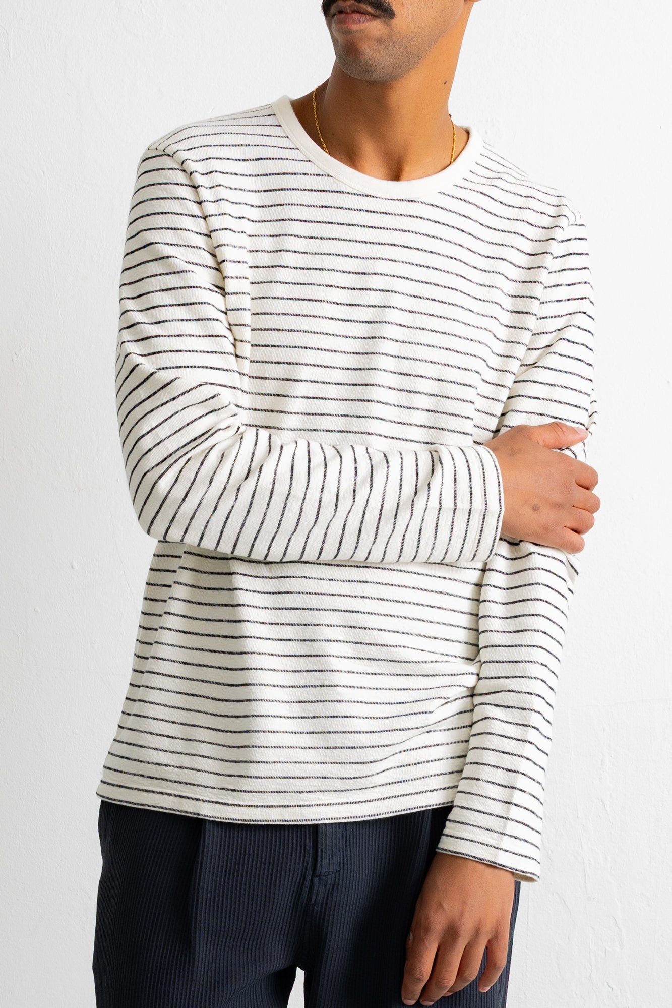 A man wearing a long-sleeve crewneck shirt with thin black horizontal stripes on an off-white base, paired with dark blue pants, against a plain white wall.