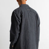 A man viewed from behind wearing a dark charcoal gray long-sleeve linen shirt and brown corduroy trousers, standing against a plain white wall.